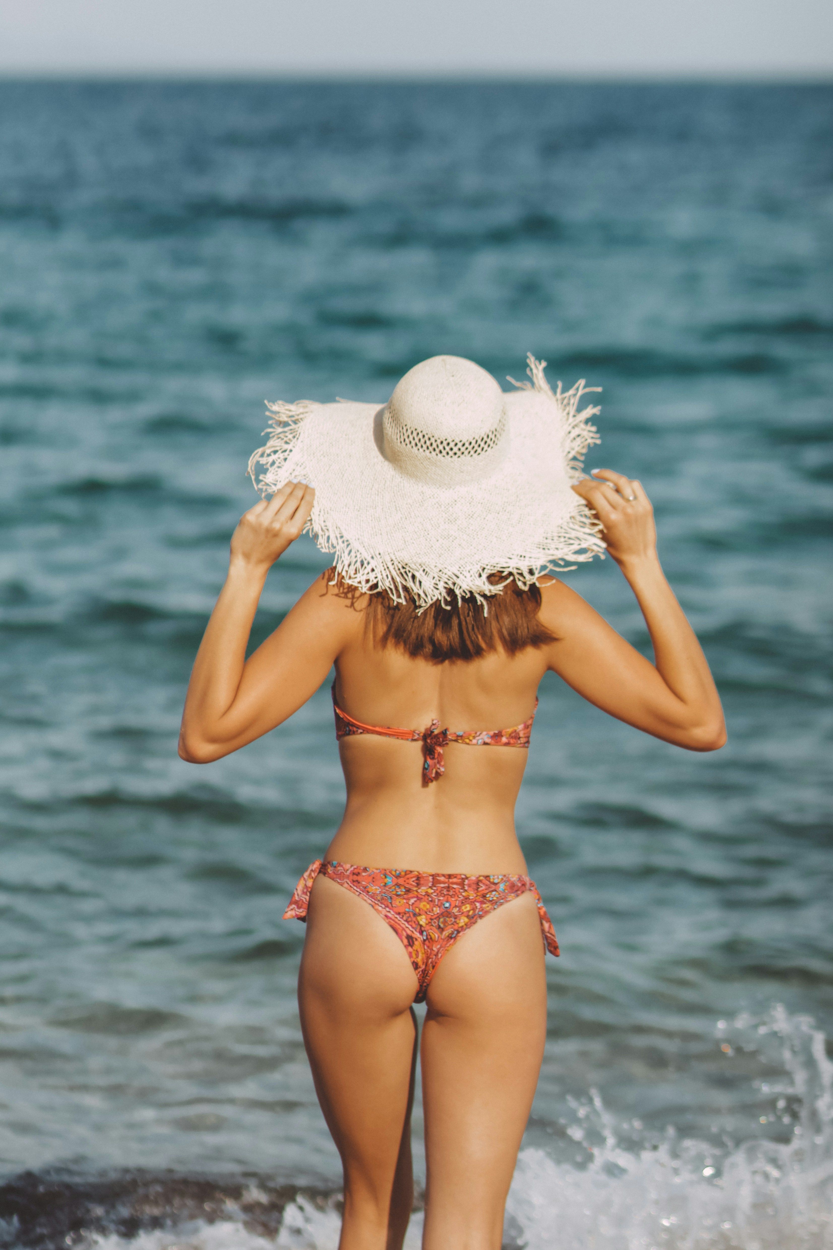 A young girl with blonde hair, seen from behind, wearing a straw hat and a bikini while looking out over a vibrant, turquoise Mediterranean cove on the Costa Blanca.