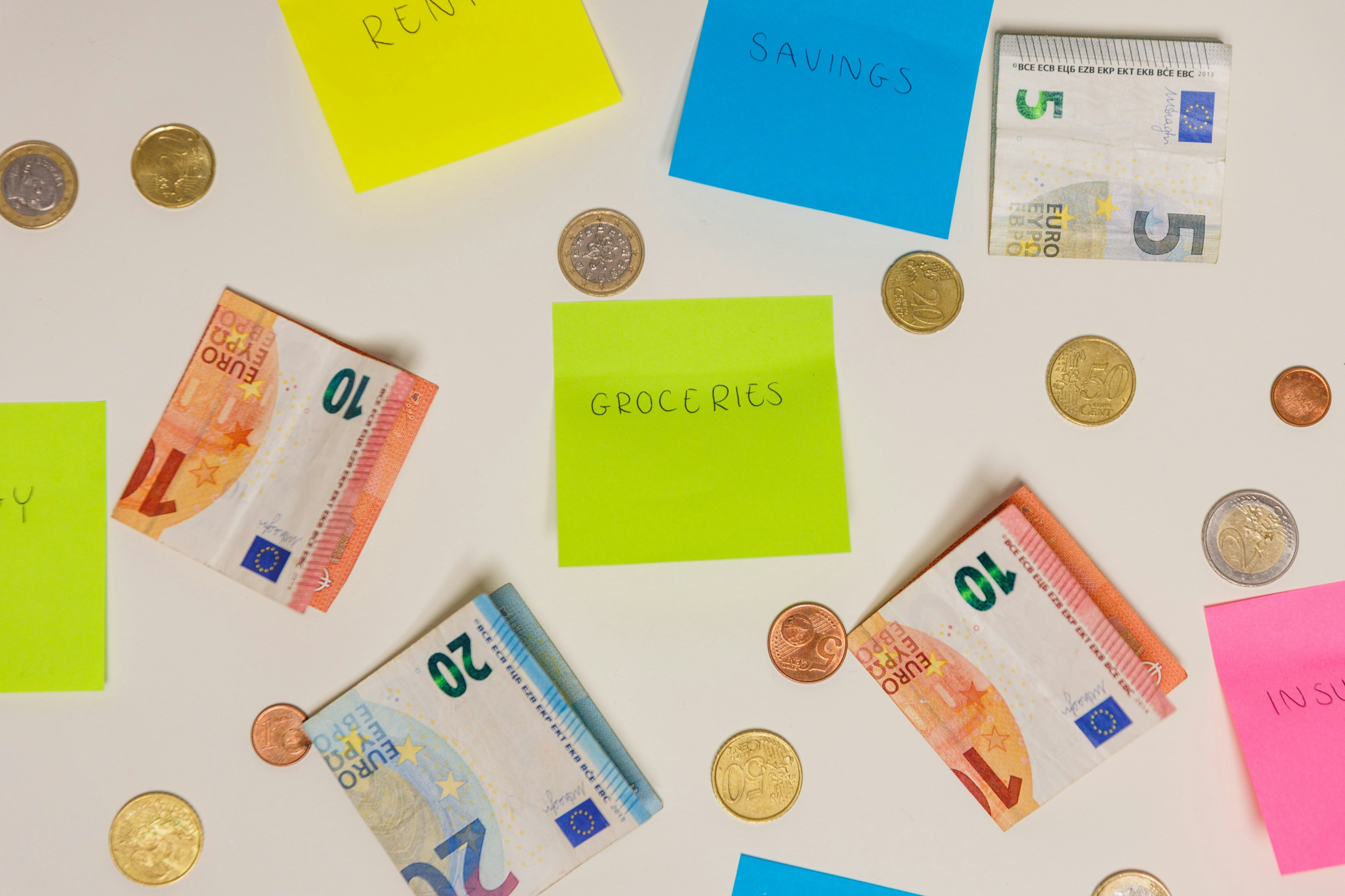 A tanble with Euro notes and coins scattered around sticky notes labeled with monthly expenses like 'Rent' and 'Groceries', representing the cost of living.